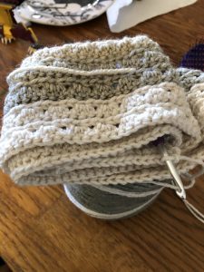 Crochet work in progress, neutral colors, sitting atop a spool of yarn. The gold crochet hook sticks out of the right bottom corner.